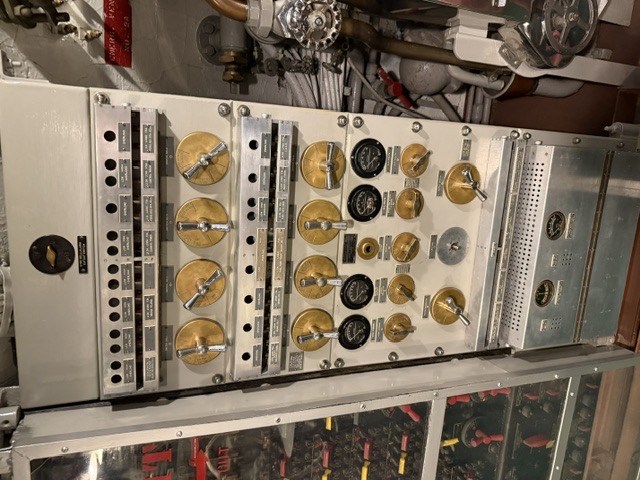 A gray steel electrical switchboard aboard USS Alabama fitted with rows of brass-handled rotary switches, labeled circuit breakers, and analog volt/amp meters, with piping running alongside on the right.