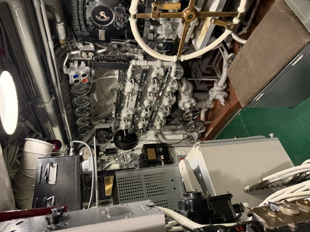 Interior view of an engine room maneuvering/throttle station aboard USS Alabama, showing valve manifolds, a brass throttle wheel, gauge panels, and white-lagged piping over a green deck.